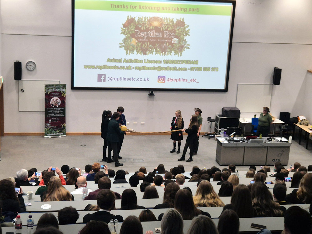 Students watching a group of volunteers holding a rope on stage while a rat runs along the rope