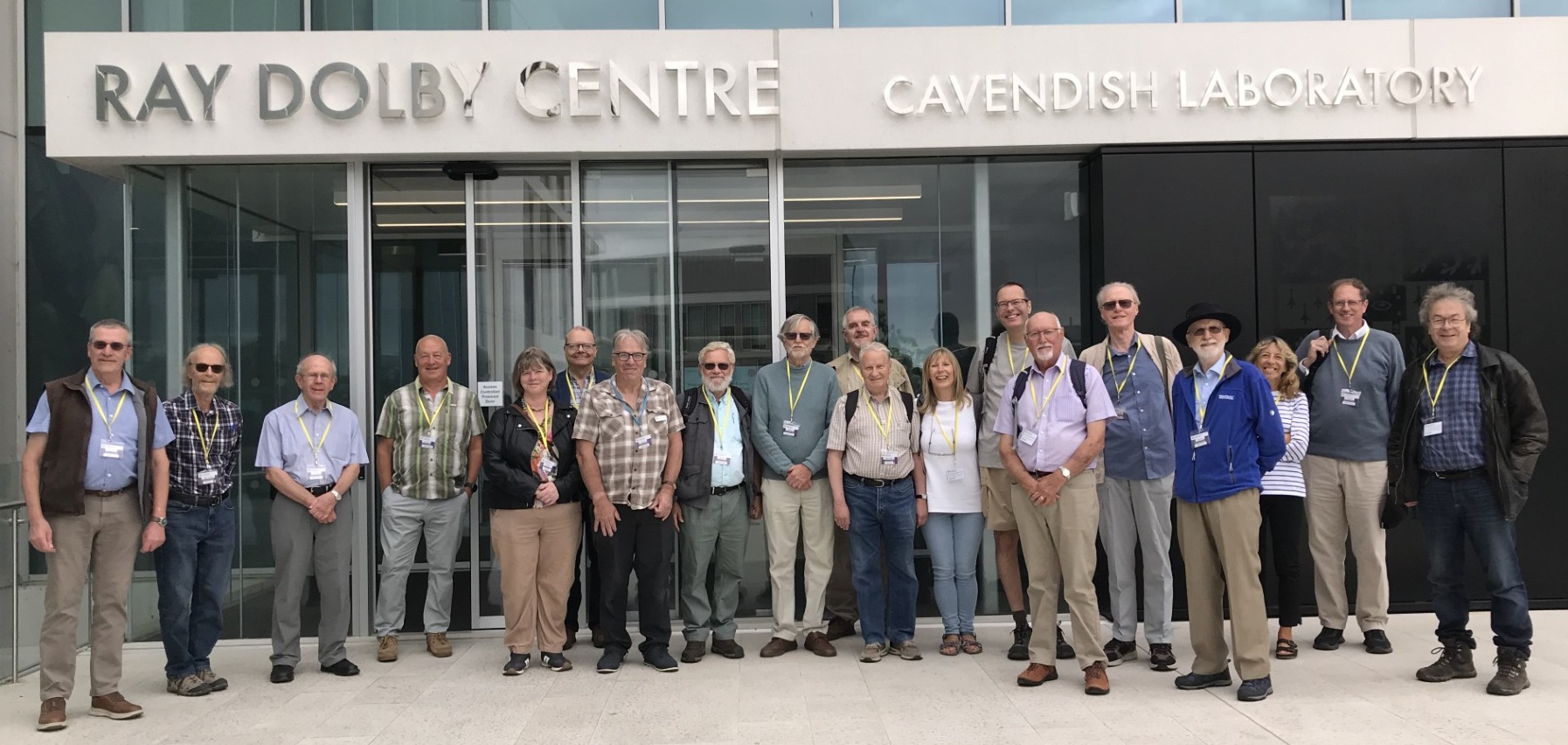 The CSES tour group smiling and standing in front of a glass-fronted building with large silver letters identifying it as the Ray Dolby Centre of the Cavendish Laboratory